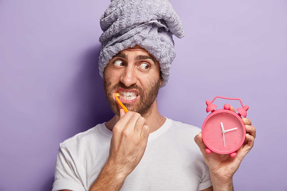 handsome-man-brushes-teeth-whitens-with-toothpaste-holds-alarm-clock-hand-awoke-late-morning-has-wrapped-towel-head-wears-casual-white-t-shirt-isolated-purple-wall-morning-routine-dental-blog